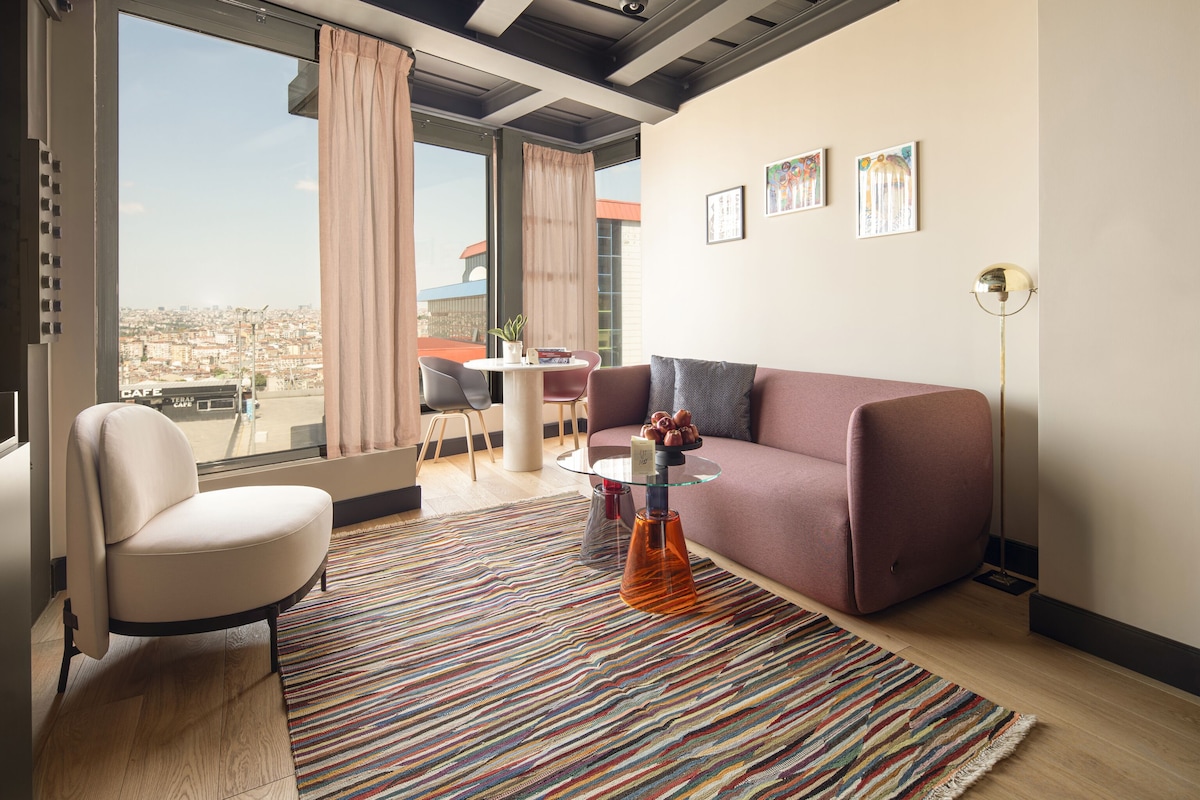 Ein modernes Wohnzimmer mit einer Couch, einem Sessel und einem Glastisch. Ein großes Fenster mit Vorhängen bietet Blick auf die Stadt, und ein farbenfroher Streifen Teppich liegt auf dem Boden.