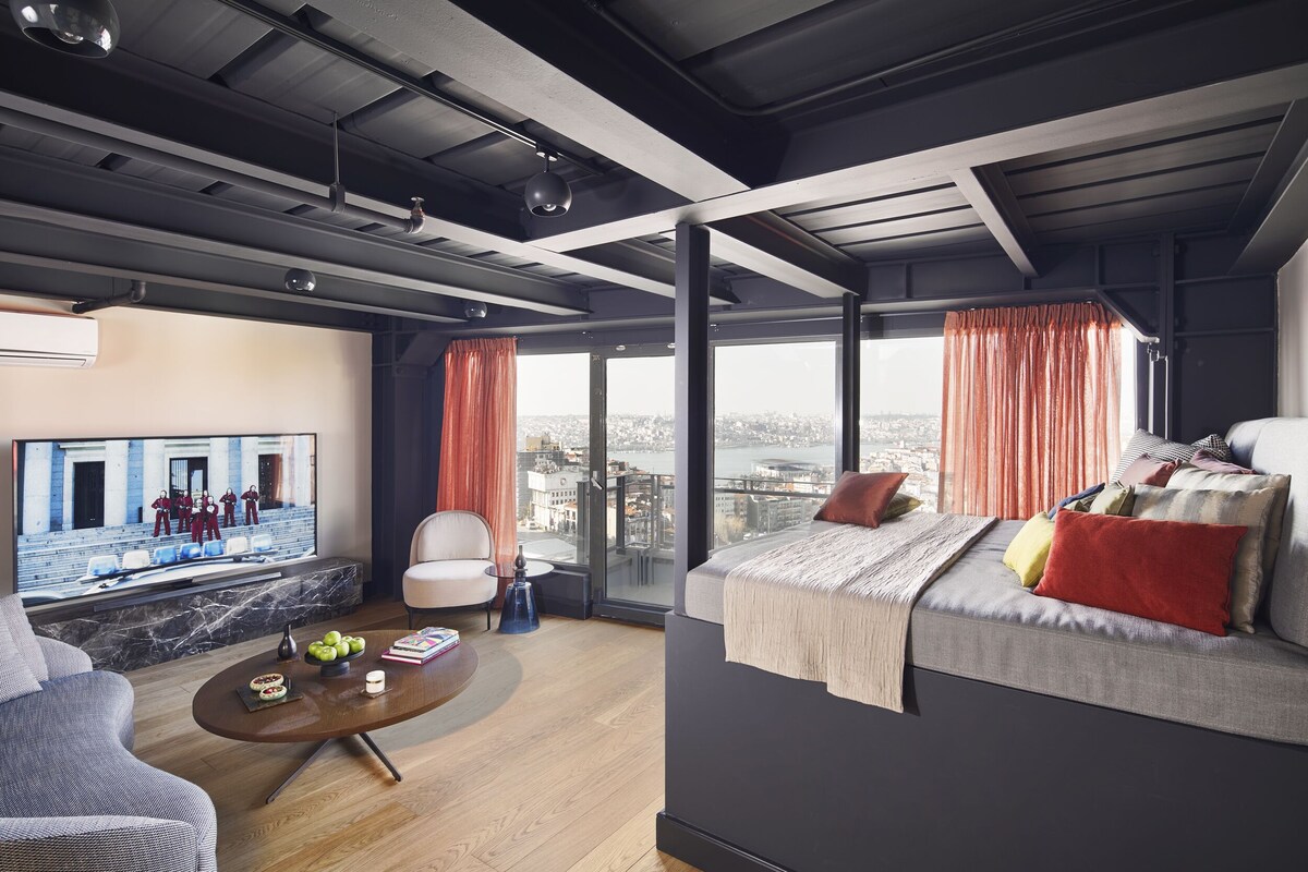 Ein modernes Hotelzimmer mit einem großen Bett, einer Sitzecke mit Couchtisch und einem Fernseher, der eine Stadtansicht zeigt.