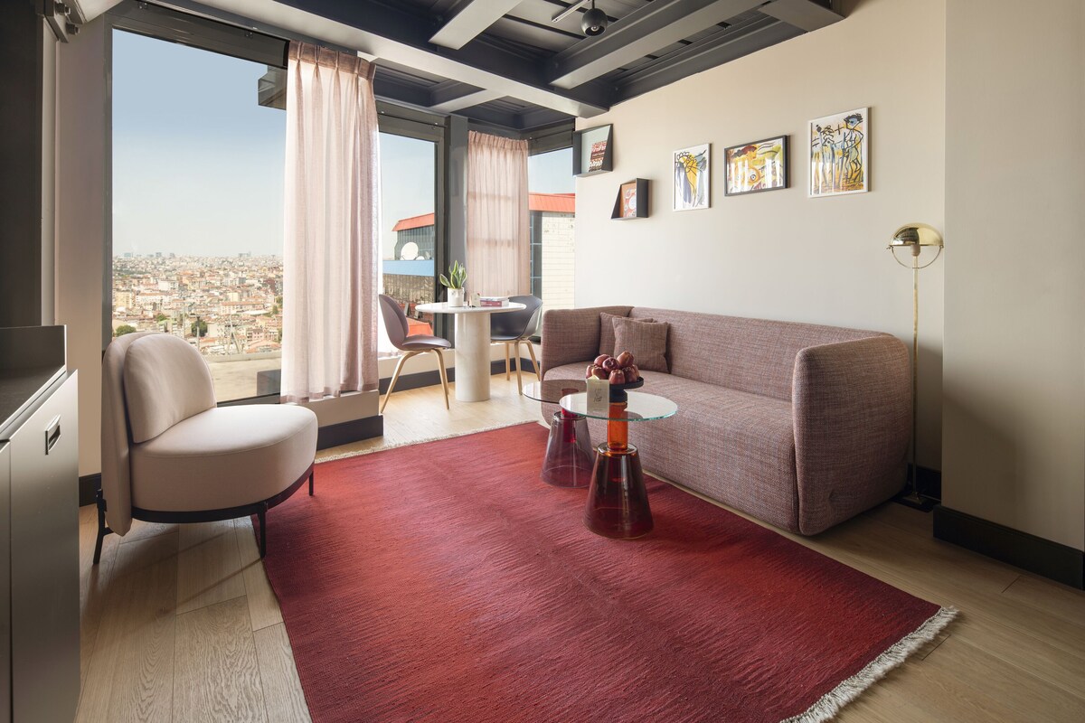 Ein modernes Wohnzimmer mit einer Couch, einem Sessel, einem kleinen Tisch und einem roten Teppich. Ein Fenster bietet Blick auf eine Stadtlandschaft.