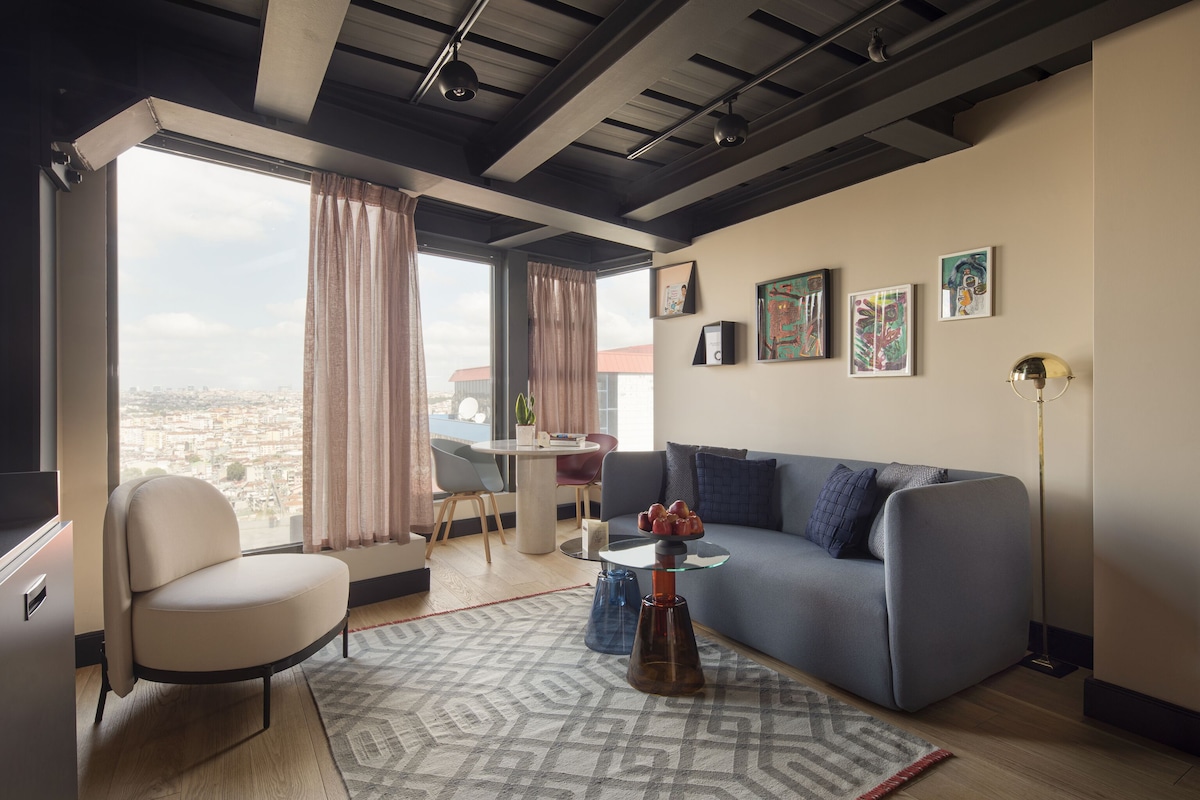 Ein modernes Wohnzimmer mit einer Couch, einem Glastisch und einem Sessel. Vor den Fenstern hängen Vorhänge, und an der Wand ist ein Kunstwerk zu sehen.