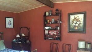 Une pièce avec un plafond en bois, un lit recouvert d’une courtepointe, une étagère contenant divers objets, dont une boîte de Coca-Cola, deux chaises, un miroir et un tableau encadré accroché au mur.