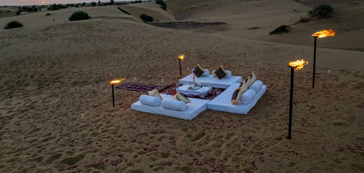 Paisagem desértica com área de estar externa luxuosa, almofadas brancas e uma mesa com manta e garrafa.