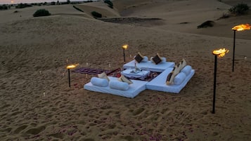 Paisagem desértica com área de estar externa luxuosa, almofadas brancas e uma mesa com manta e garrafa.