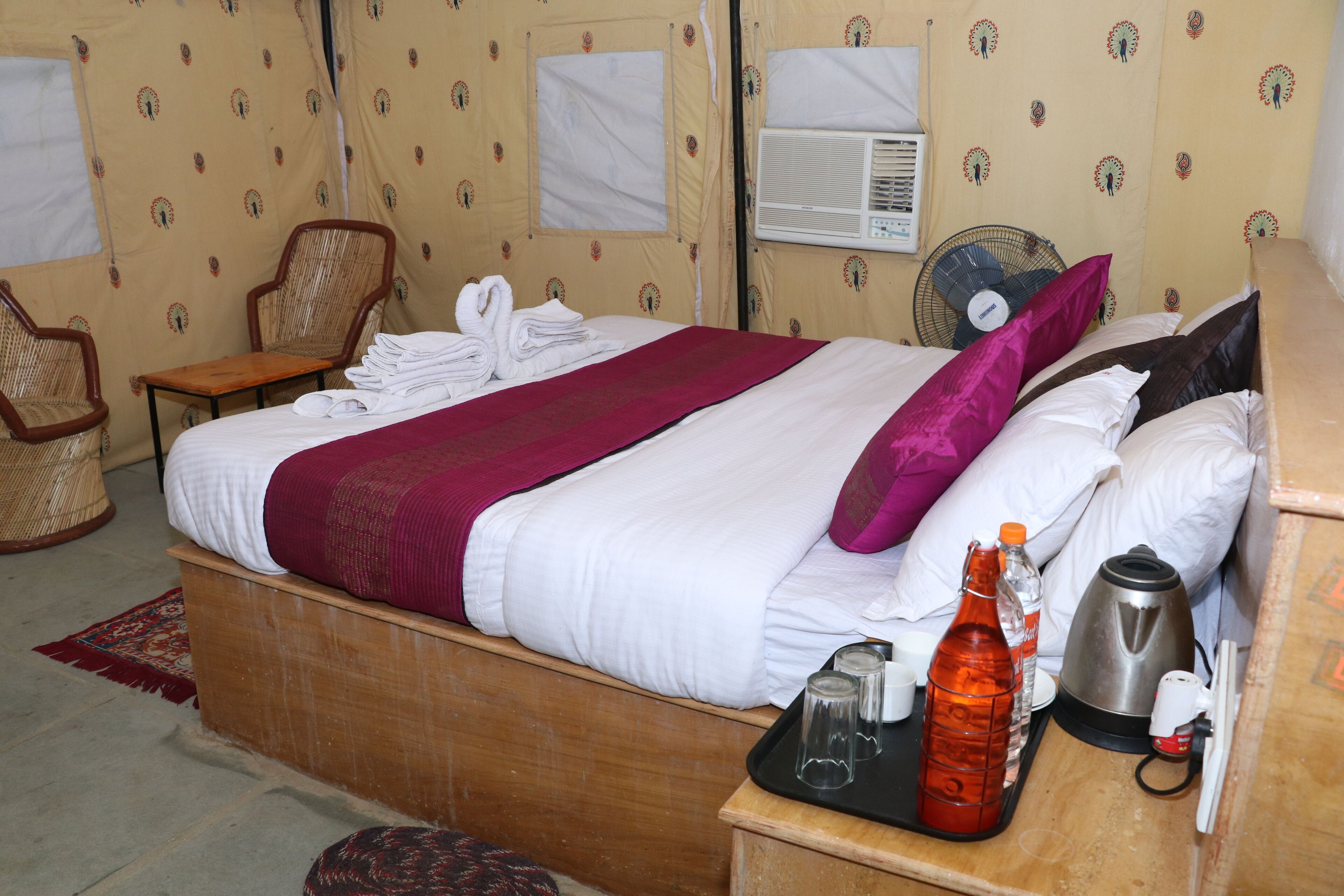 Ein Zimmer mit einem Bett, einem Nachttisch aus Holz, einem Ventilator und einem Fenster mit Vorhängen.
