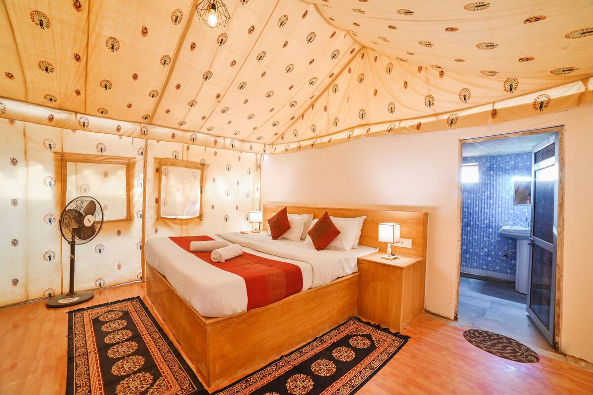 Un dormitorio con una cama de madera, dos mesitas de noche con lámparas, un ventilador de pie y una alfombra estampada.
