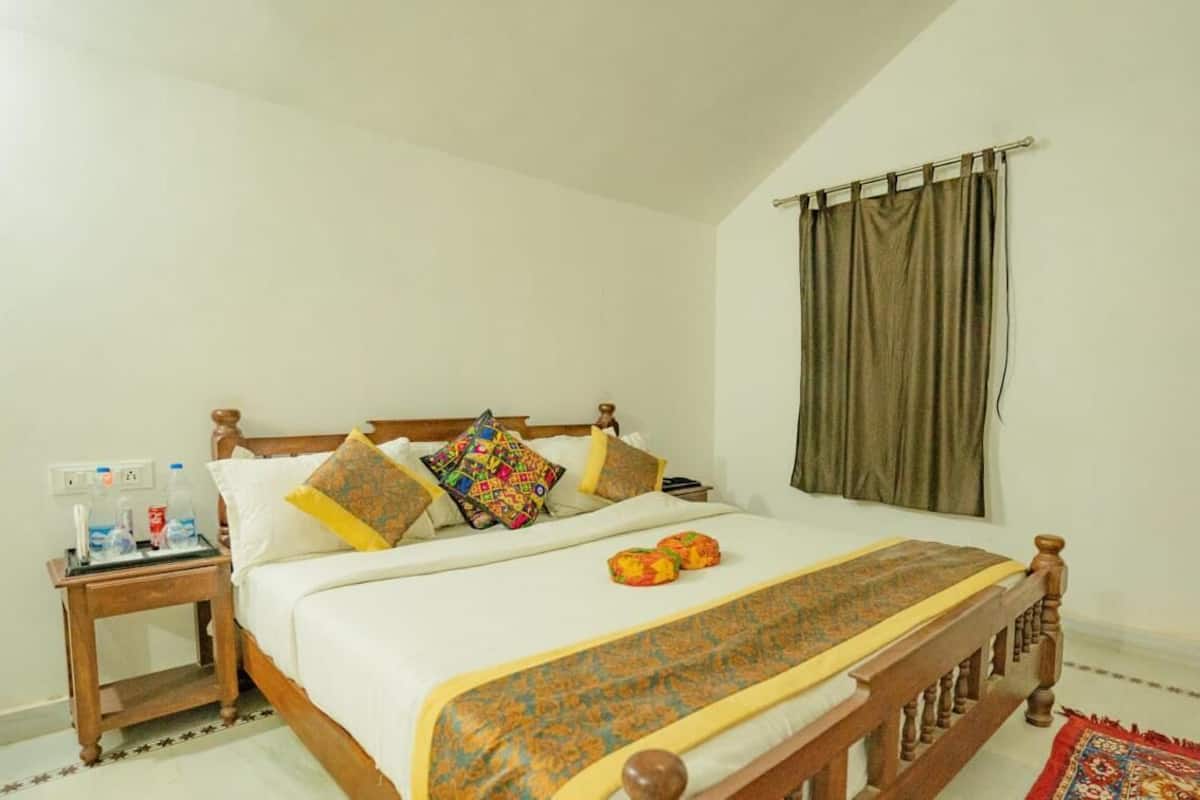 Un dormitorio con una cama de madera, ropa de cama blanca, almohadas coloridas, una mesita de noche con botellas de agua y una cortina en la ventana.