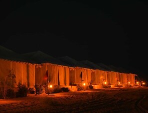A large, illuminated building with multiple windows and a flat roof, surrounded by sparse vegetation. - Shama Desert Camp (Jaisalmer)