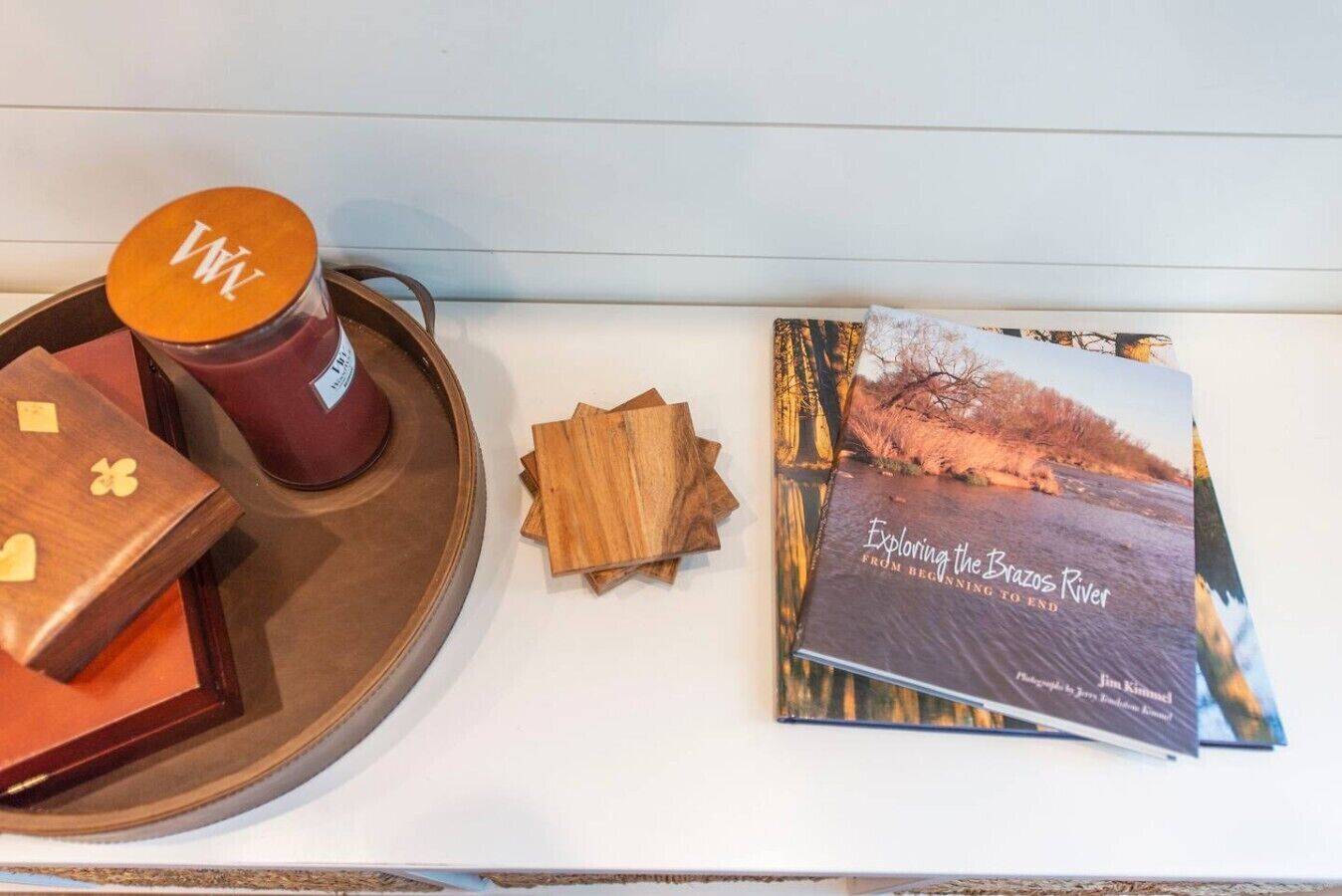 Una bandeja con una vela, posavasos de madera, un libro titulado 'Exploring the Brazos River' y una caja de madera con diseño de corazón.
