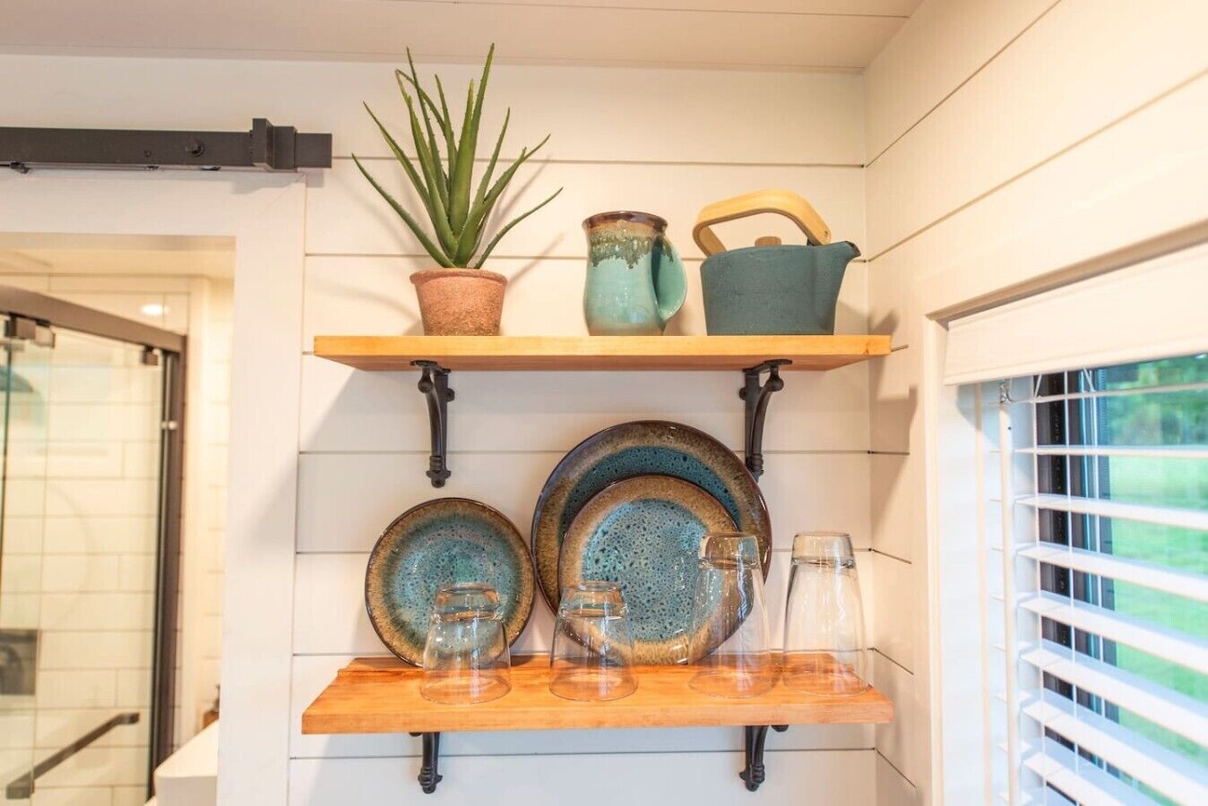 A kitchen shelf with a potted plant, a pitcher, a teapot, and two plates with glasses.