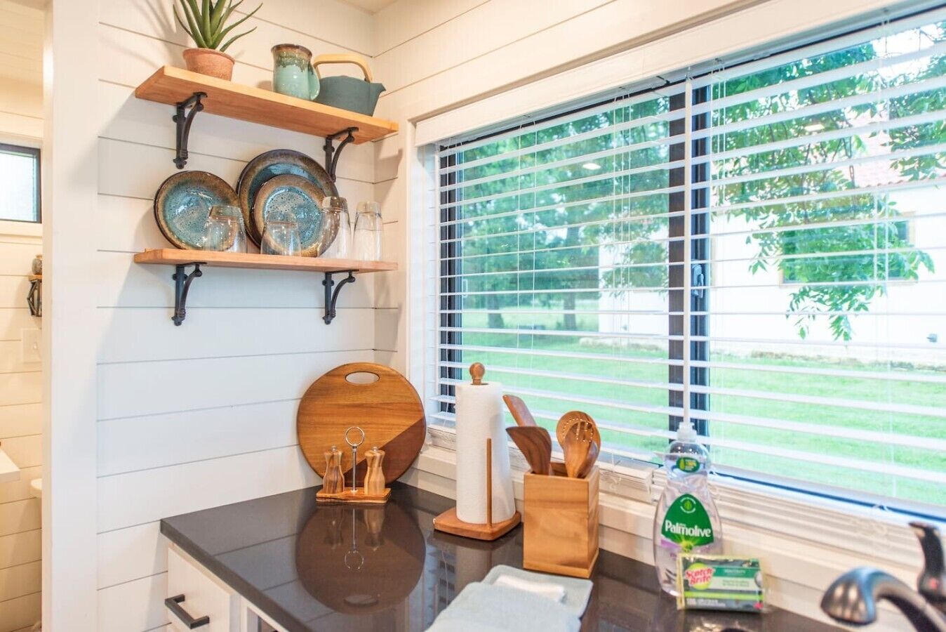 A kitchen with wooden shelves, a black countertop, and a window with blinds.