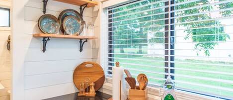 A kitchen with wooden shelves, a black countertop, and a window with blinds.