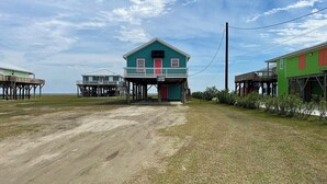 A coastal house with a porch and a small deck, surrounded by grassy areas and other buildings. - Sassy Seagull Beachfront and Sunrise (Grand Isle)