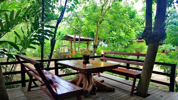 Eine hölzerne Terrasse im Freien mit einem Tisch in der Mitte und Bänken, umgeben von üppigem Grün und einem Gazebo im Hintergrund.