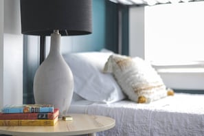 Una cama con ropa blanca, una almohada blanca, un perro blanco pequeño y una mesita redonda con libros y una lámpara.