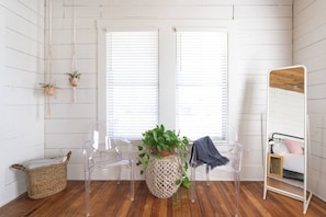 Una habitación con paredes de madera blanca, suelo de madera, dos ventanas con persianas, una silla transparente, una cesta tejida, una planta en maceta y un espejo sobre un atril.
