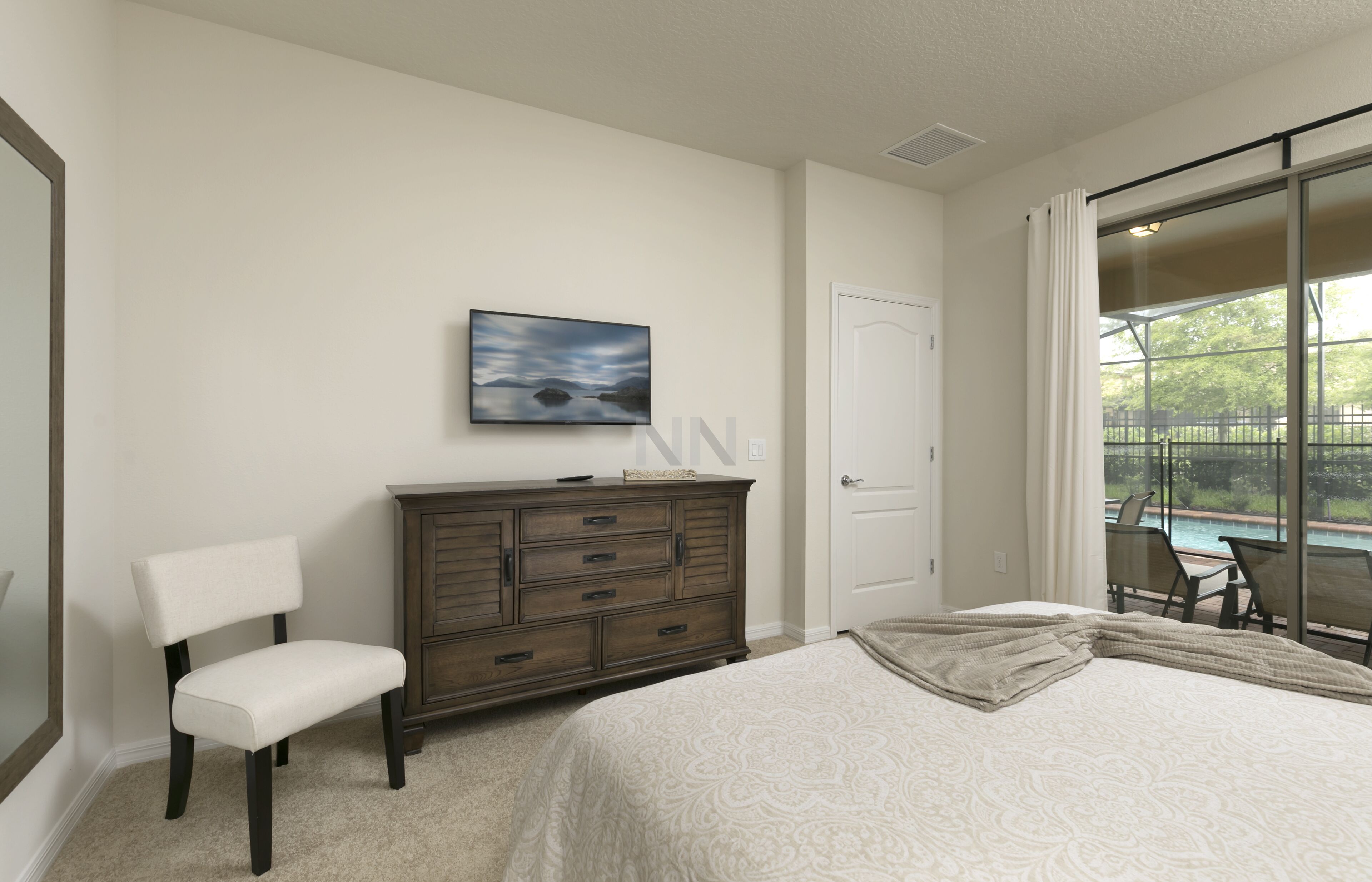 A bedroom with a bed, a dresser, a chair, a painting, and a view of a pool through a sliding glass door.