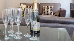Et sofabord af glas med champagneglas og en flaske Brut vin i en moderne stue med en grå sofa og mønstrede puder.