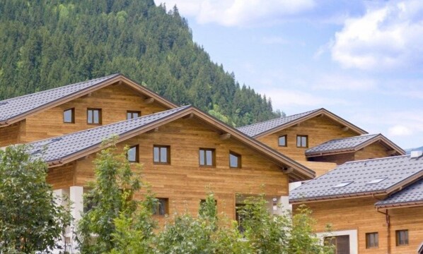 A row of wooden cabins with pitched roofs, set against a backdrop of a forested mountain.