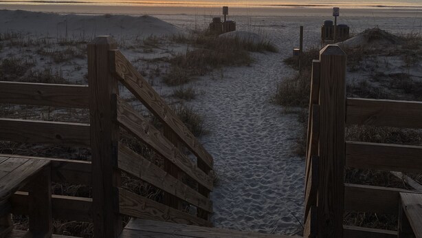 Ein hölzerner Steg führt zu einem Strand, im Hintergrund ein Sonnenuntergang.
