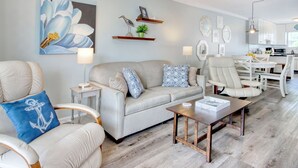 Ein Wohnzimmer mit Sofa, Sesseln, einem Couchtisch und einem Essbereich mit Stühlen und Tisch. Im Hintergrund ist eine Küche zu sehen.