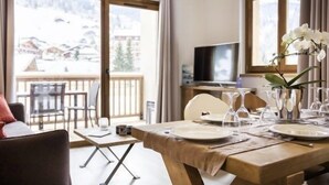 A dining area with a wooden table set for four, a TV in the background, and a balcony with a view of a snowy landscape. - Vos Prochaines Vacances de Ski | Appart Confortable avec Cuisine (Arêches Beaufort)