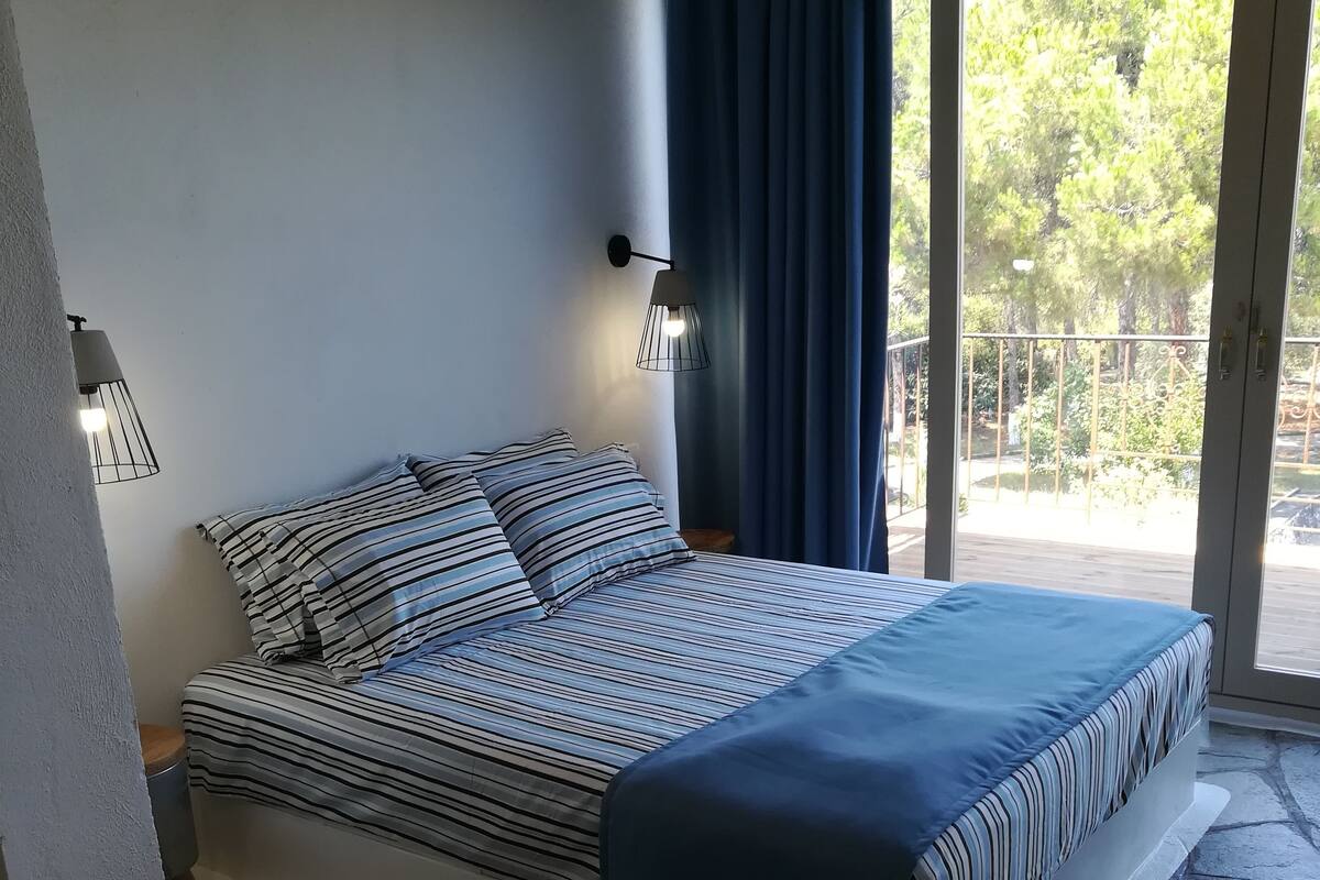 A bed with striped bedding, a nightstand, a wall-mounted lamp, and a view of an outdoor balcony with a railing.