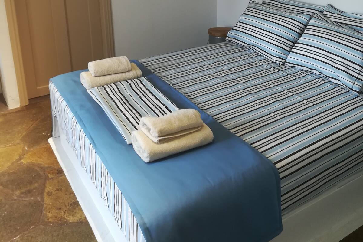 A neatly made bed with striped bedding and towels, a wooden door, and a wall-mounted lamp.