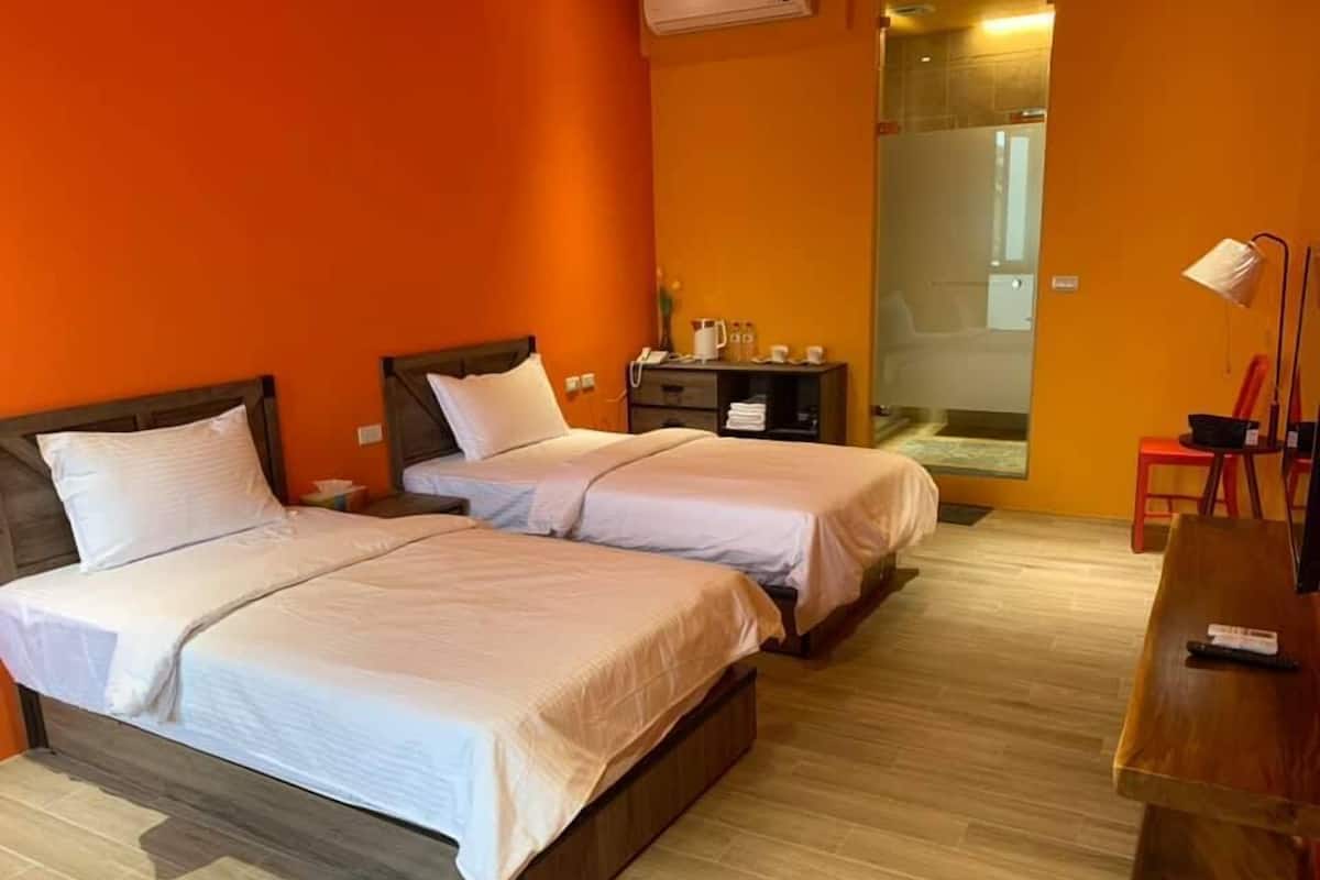 A hotel room with two beds, a wooden floor, a desk with a lamp, and a bathroom visible through a glass door.