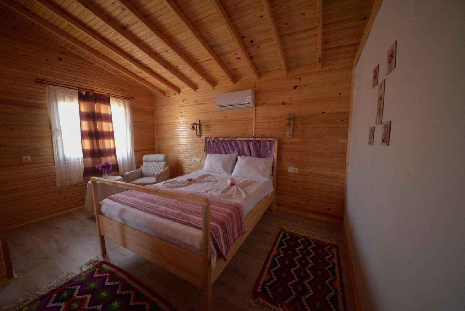 Un dormitorio de cabaña de madera con una cama, una silla, una mesita de noche, una ventana con cortinas y un aire acondicionado de pared.