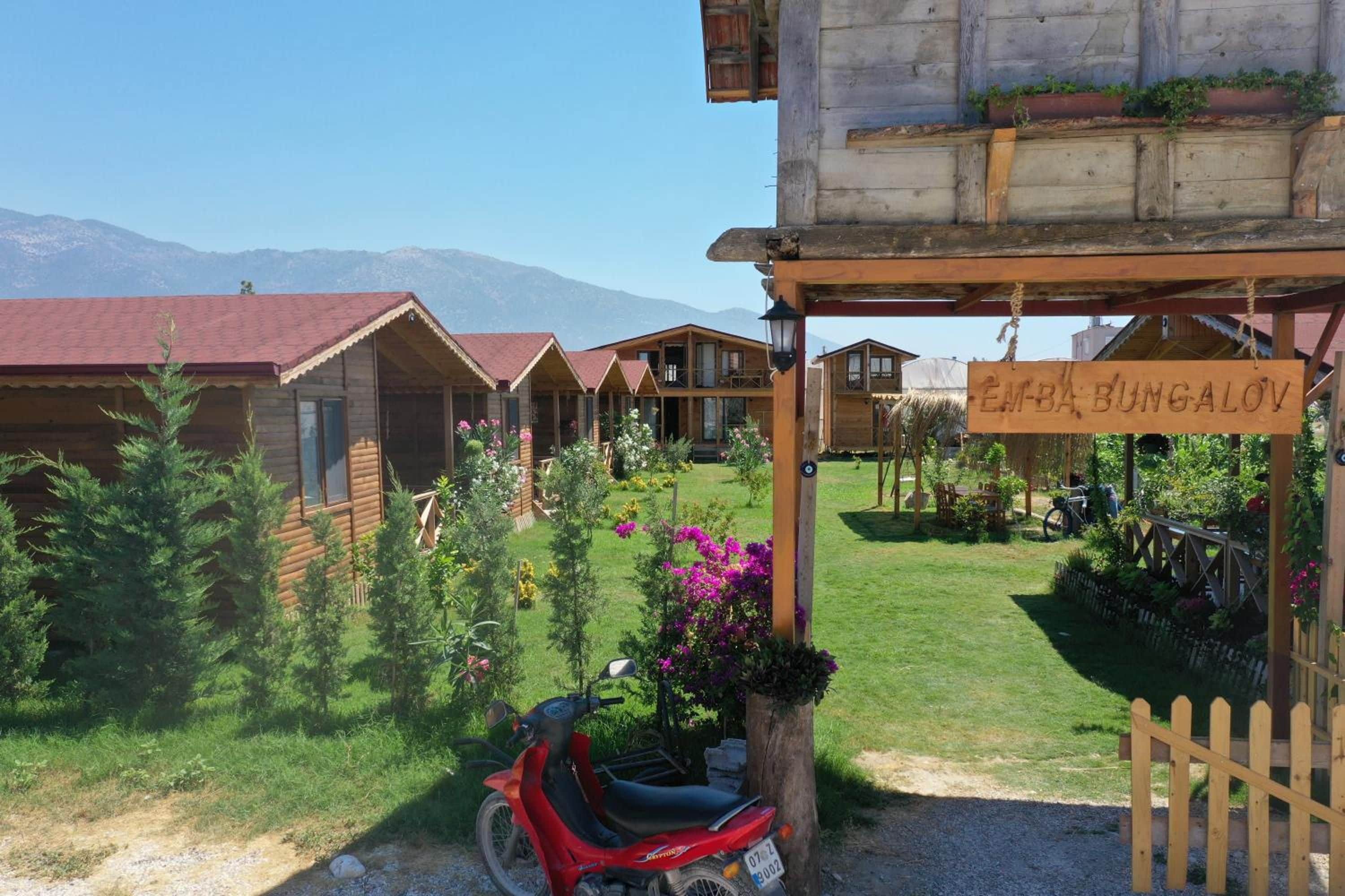 Una hilera de bungalows de madera con un letrero que dice 'EMBA BUNGALOV' en un jardín, con montañas al fondo.