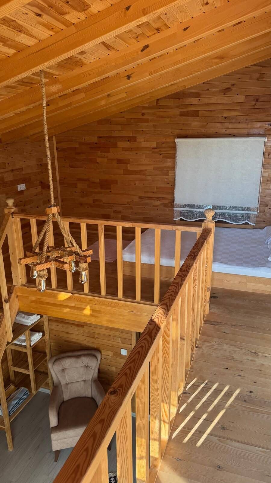 A wooden interior with a balcony, a chair, a hanging light fixture, and a window with a curtain.