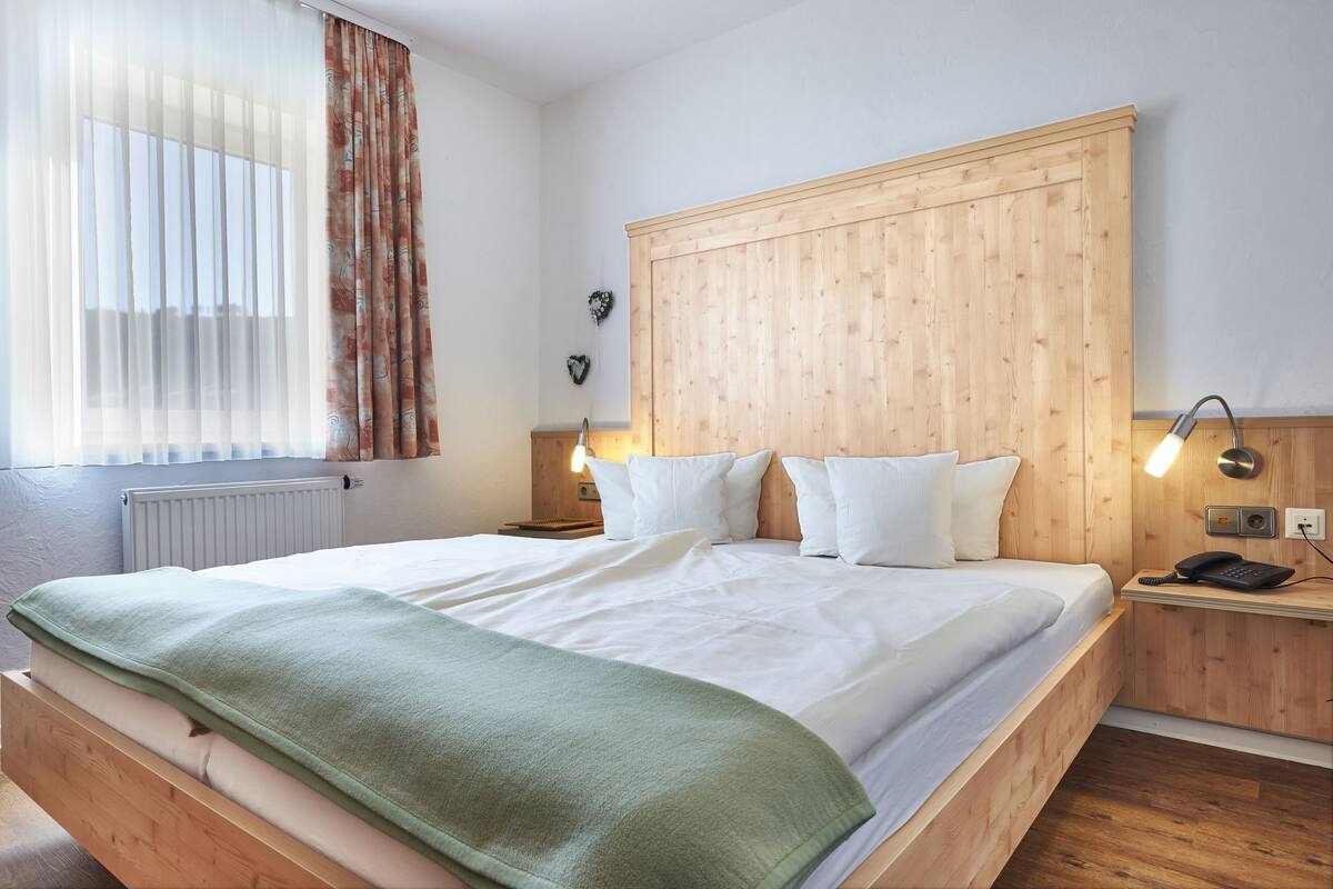 Un lit bien fait, avec du linge de lit blanc et un plaid vert, une tête de lit en bois, des lampes de chevet et une table de chevet en bois sur laquelle se trouve un téléphone.