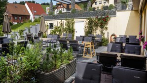 Un patio al aire libre con varias mesas y sillas, rodeado de follaje y vistas a edificios residenciales.