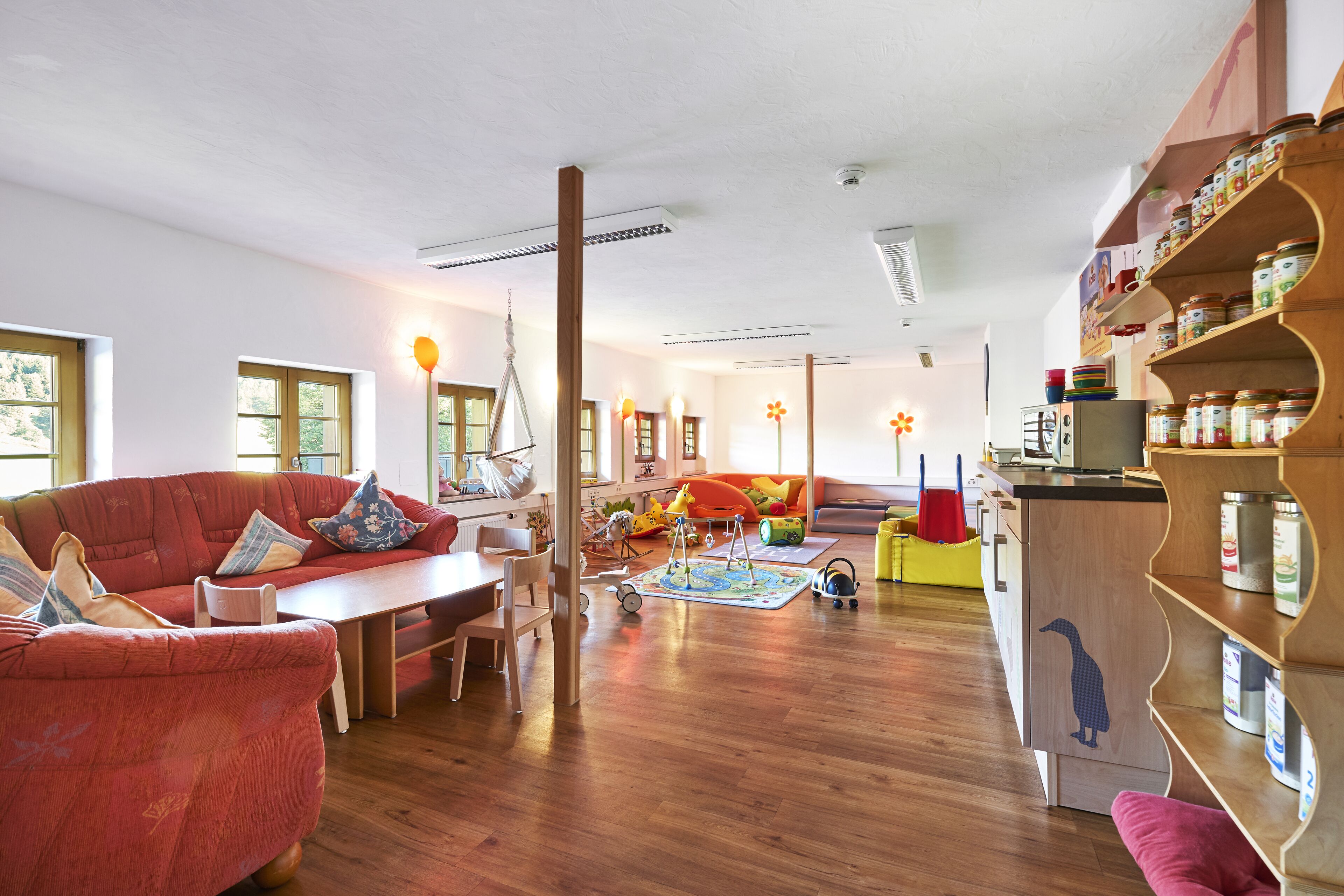 A spacious living room with a wooden floor, a red sofa, a wooden coffee table, and a bookshelf filled with jars.