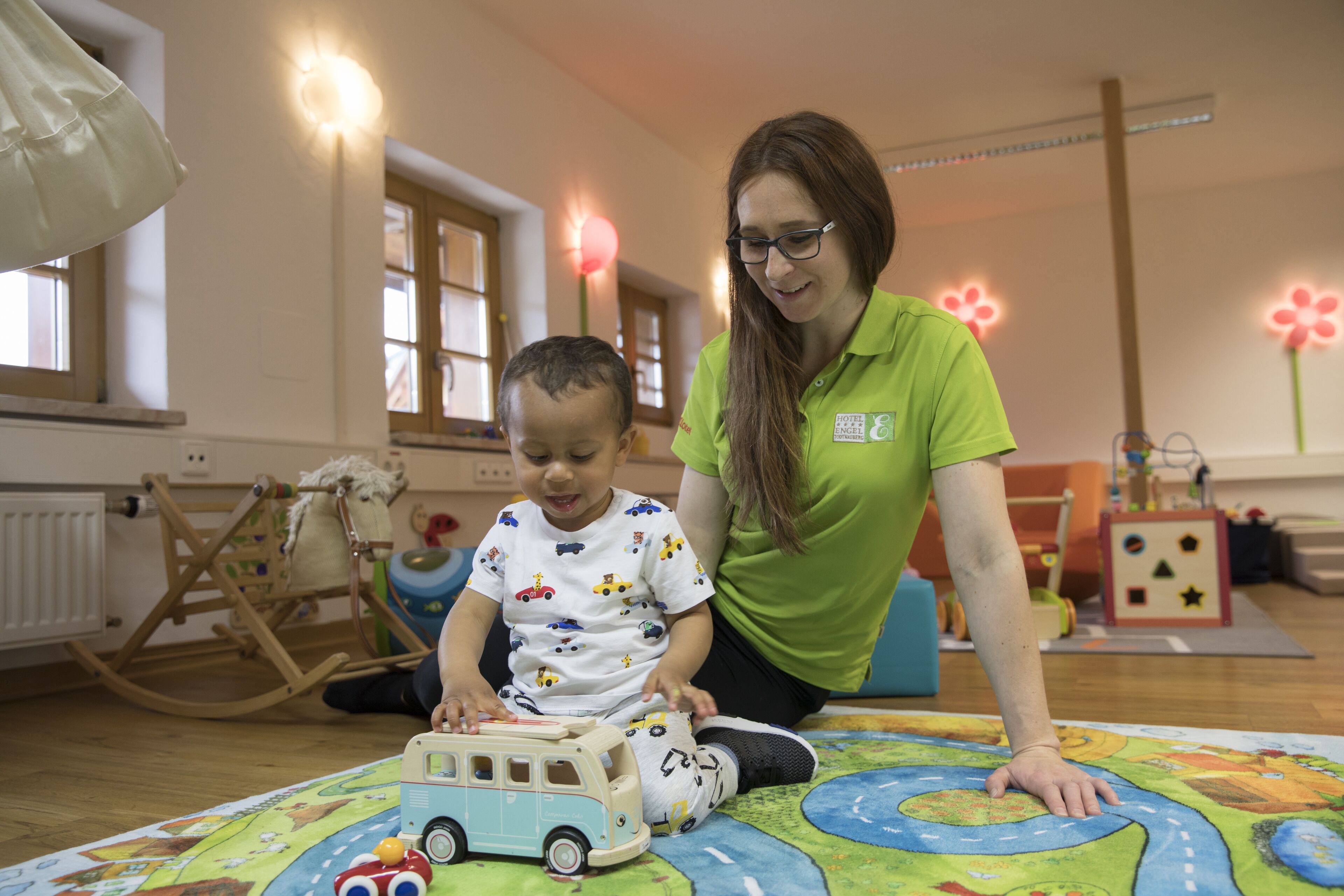 Un enfant joue avec un camion jouet sur un tapis de jeu coloré, dans une pièce dont le sol est en bois, qui contient un fauteuil à bascule et une fenêtre décorée de fleurs.