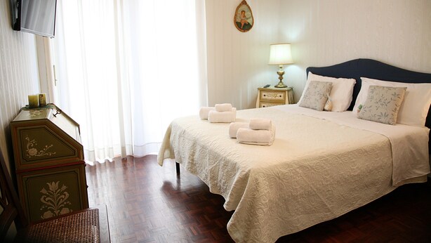Una cama bien hecha con almohadas y toallas, una mesita de noche con lámpara, un televisor montado en la pared y un aparador de madera.