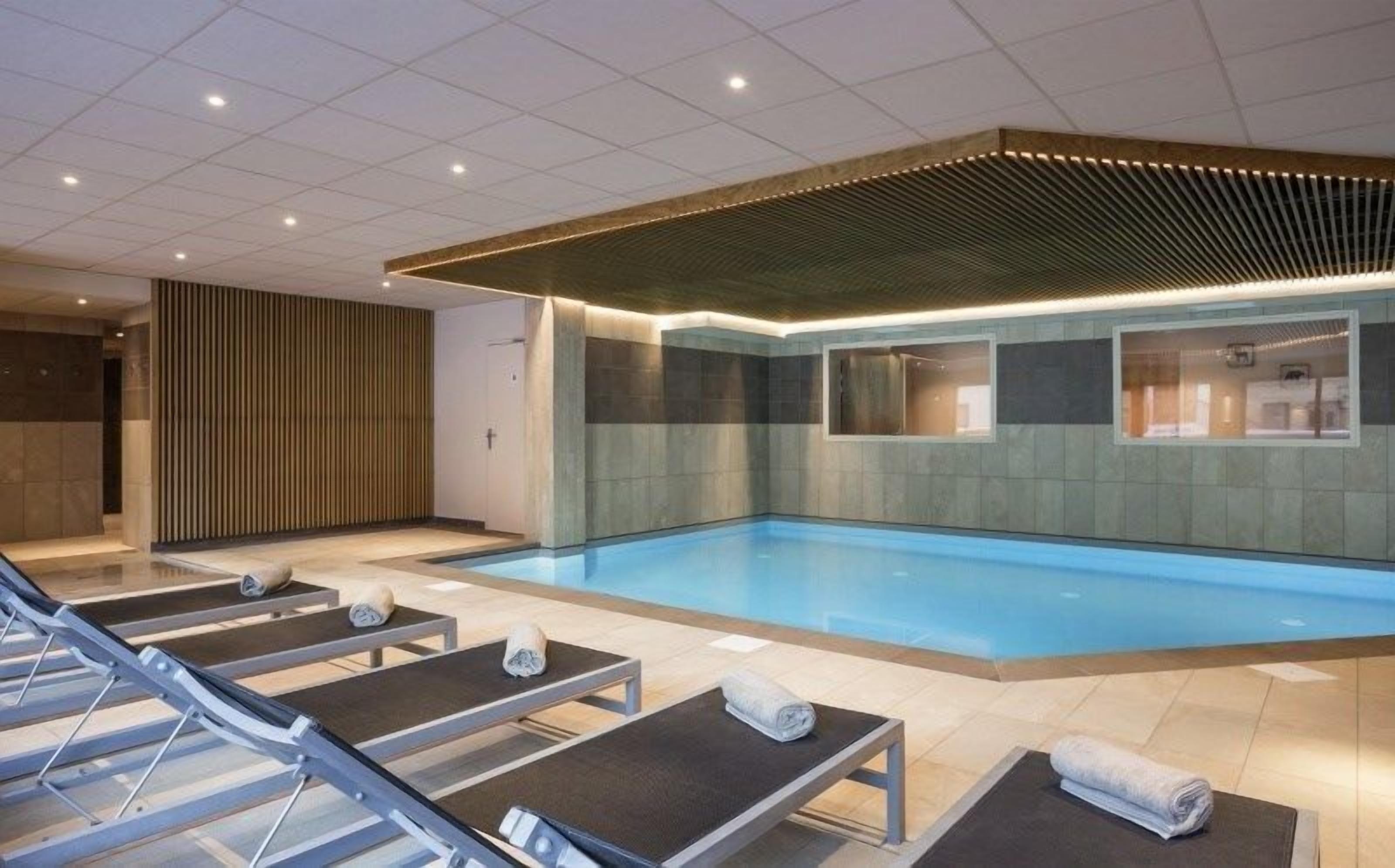Ein modernes Hallenbad mit Fliesenboden, einer Holzdeckenverkleidung und mehreren Liegestühlen mit Handtüchern.