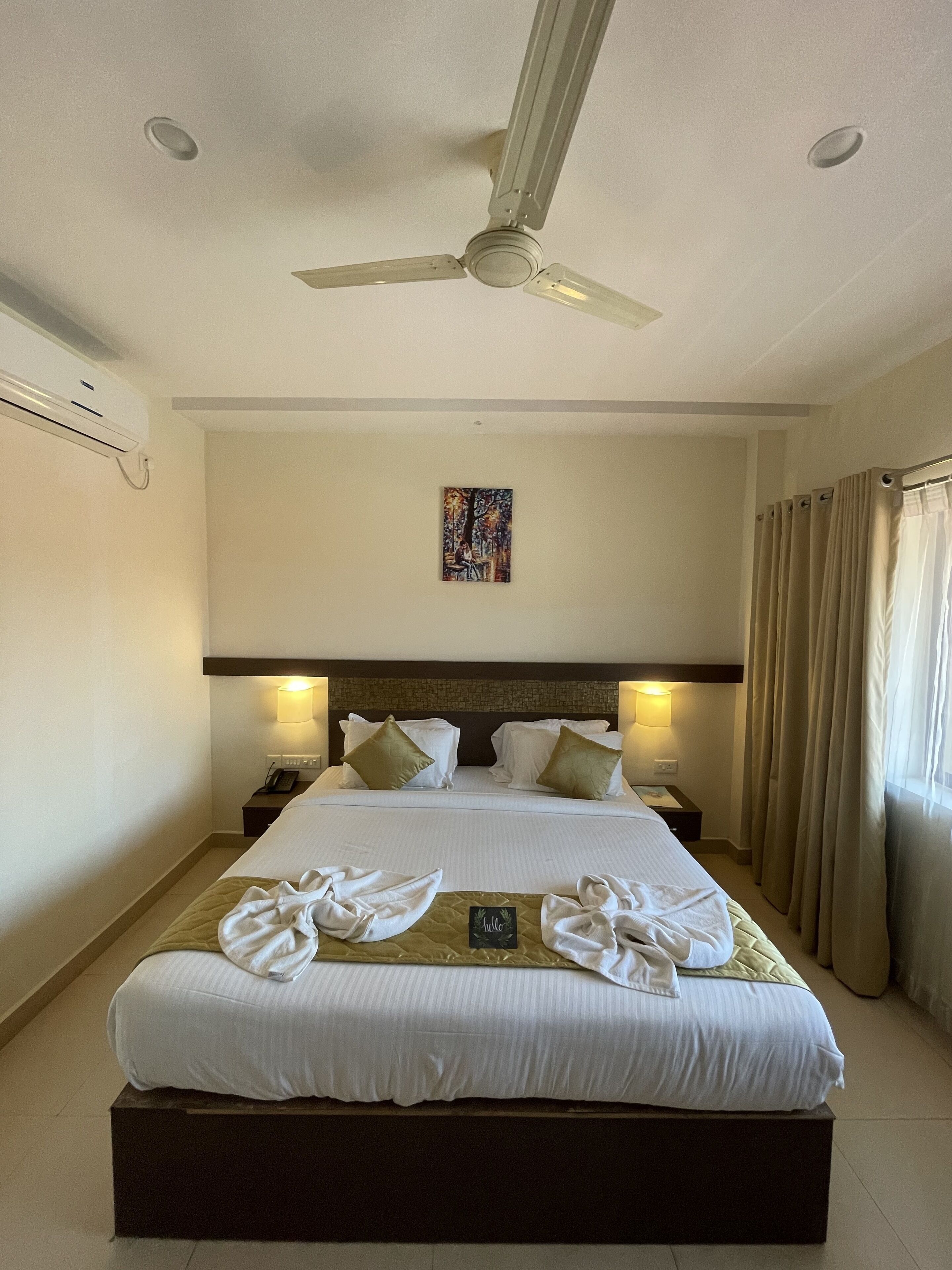 Habitación de hotel con una cama grande, un ventilador de techo, un cuadro en la pared y una ventana con cortinas.