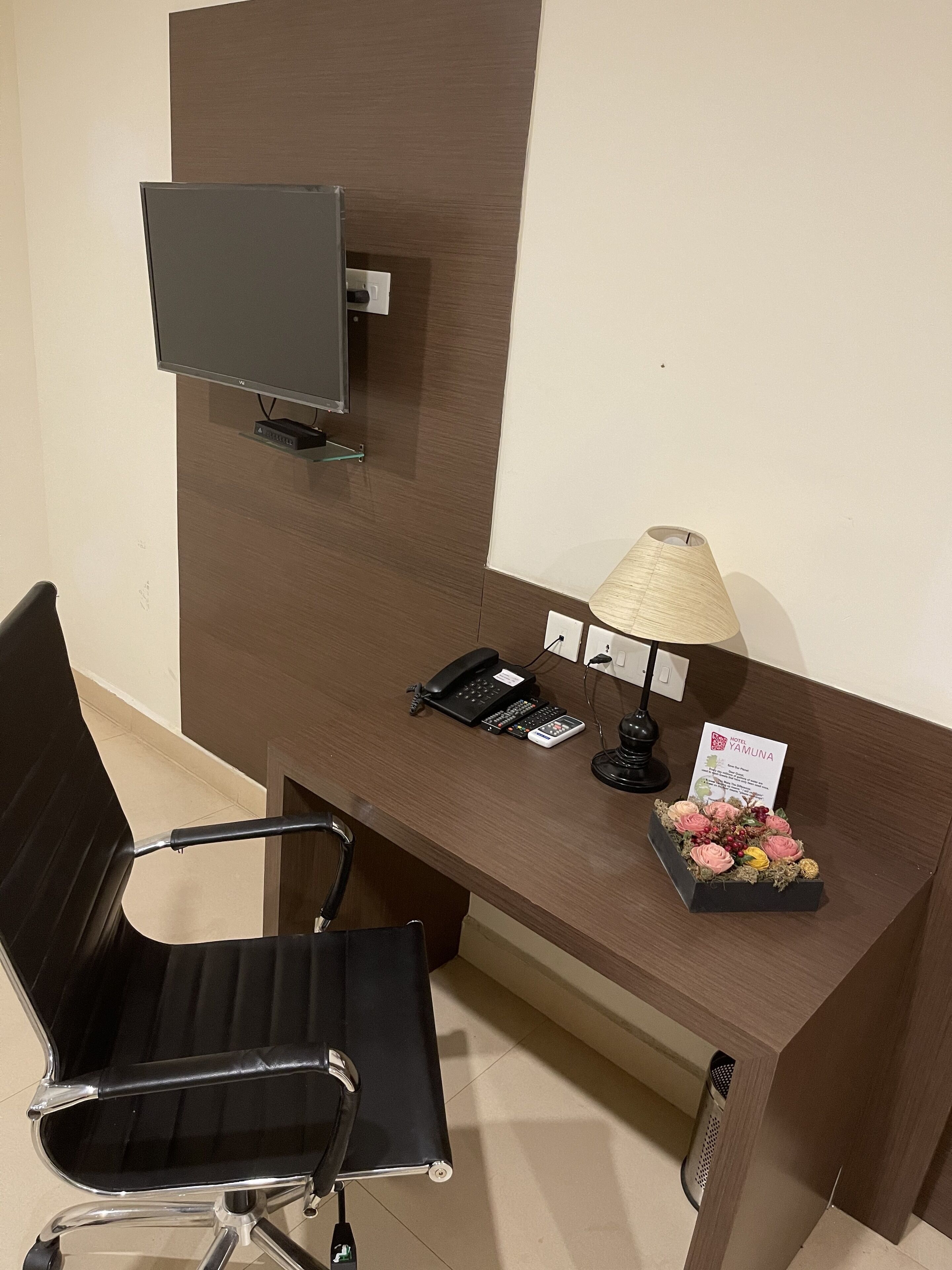 Quarto de hotel com escrivaninha de madeira, cadeira de escritório preta, televisão de tela plana na parede, luminária de mesa, telefone e um pequeno arranjo de flores.