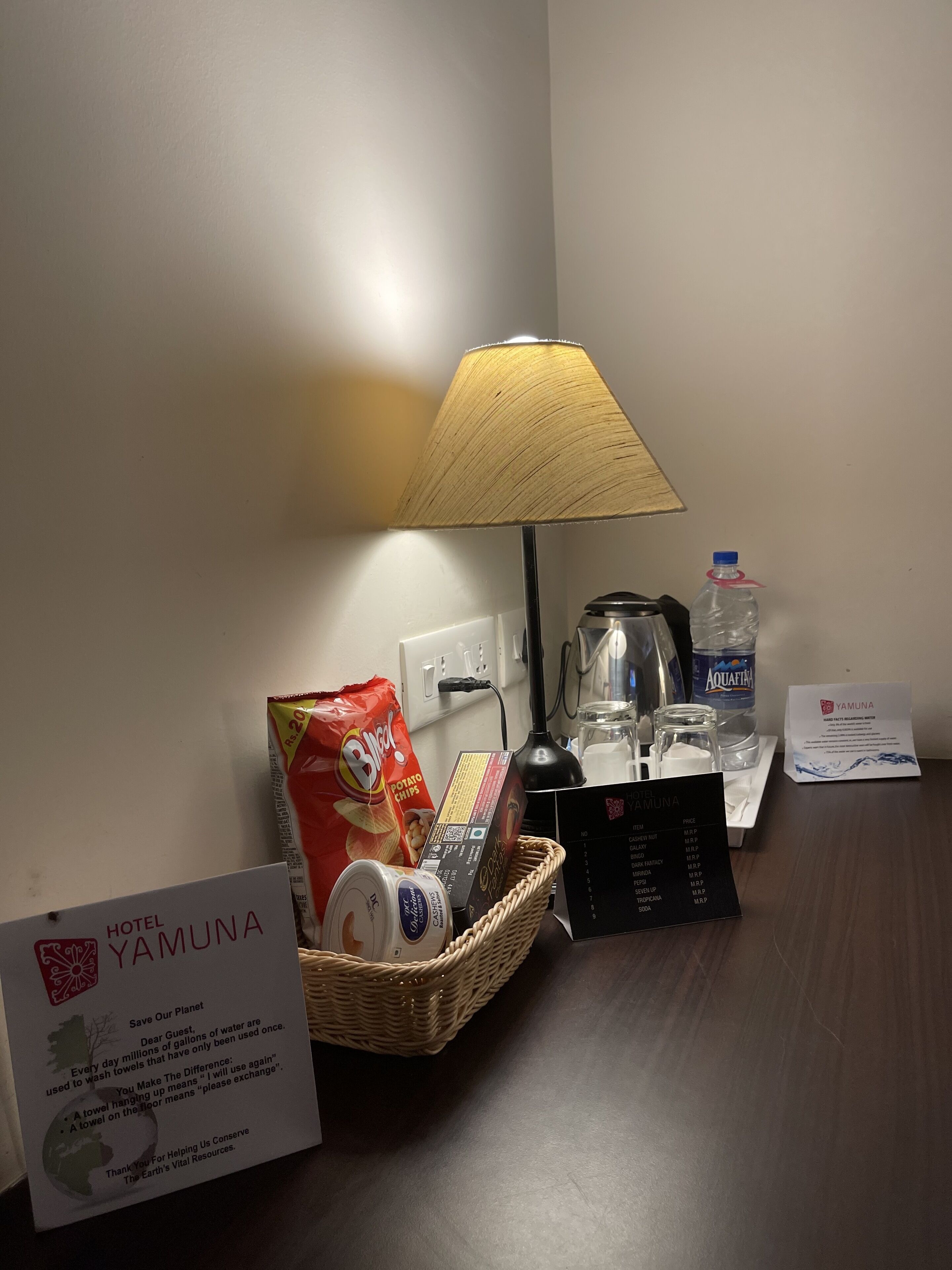 Habitación de hotel con una mesa dispuesta con aperitivos, una botella de agua, una lámpara y una toalla colgada en la pared.