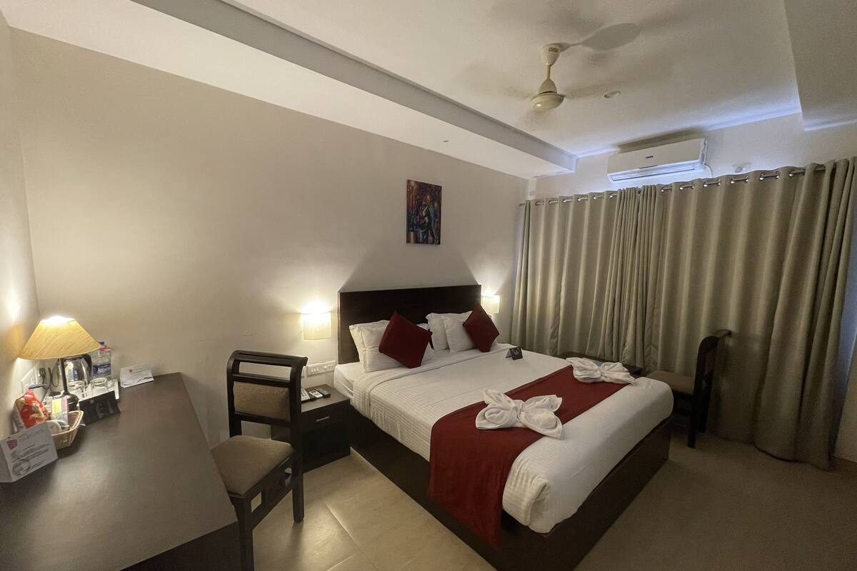 A hotel room with a bed, a desk, a chair, a lamp, a ceiling fan, and a window with curtains.