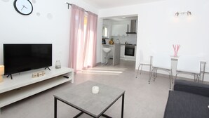 Ein modernes Wohnzimmer mit Flachbildfernseher, Couchtisch und Sofa. Im Hintergrund ist eine Küche mit weißen Schränken und Edelstahlgeräten zu sehen.