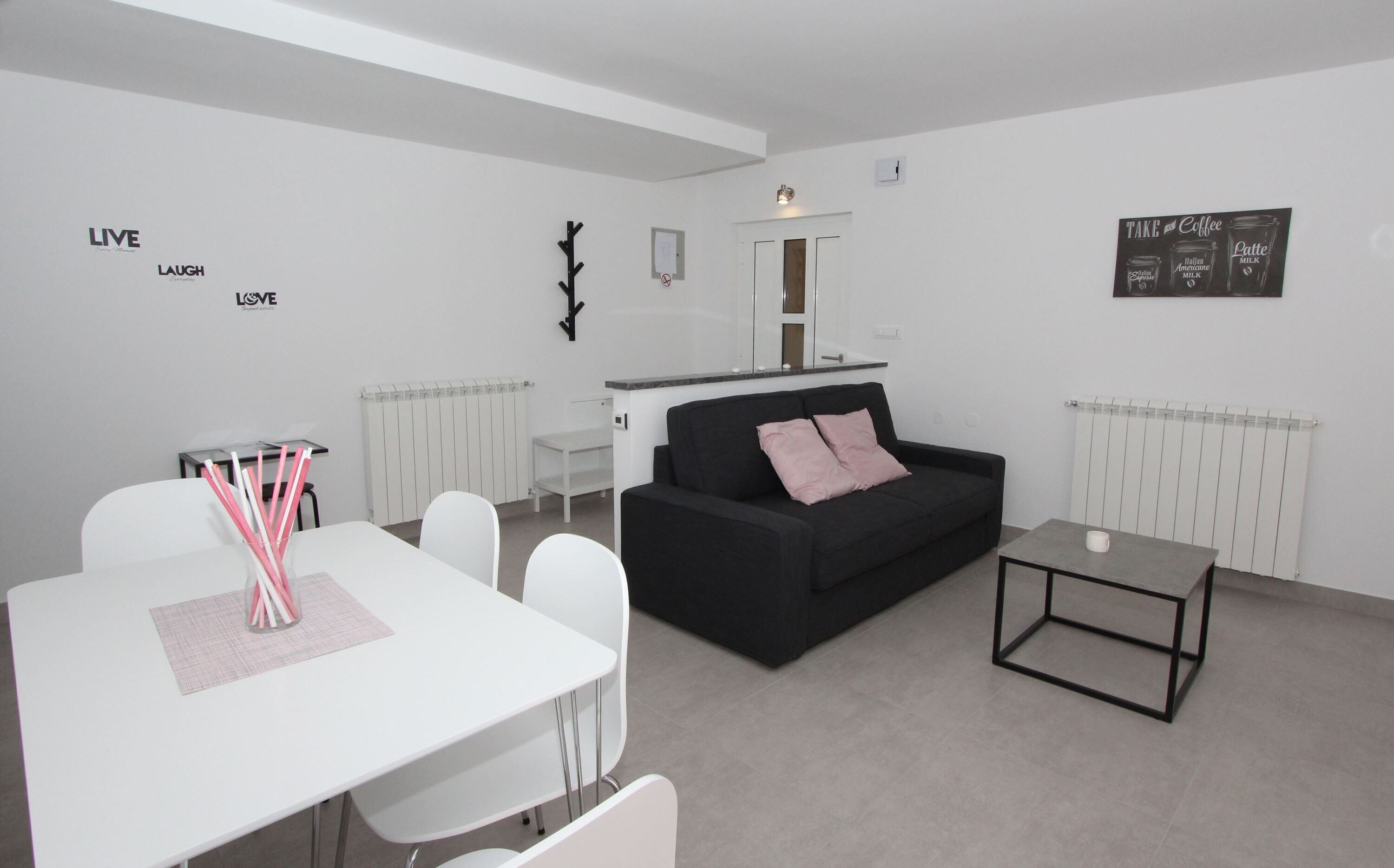 A modern living room with a black sofa, a white dining table with chairs, and a small coffee table. There are wall decorations and a radiator.