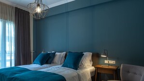 A neatly made bed with white and teal bedding, a bedside table with a lamp, and a chair in a room with teal walls and a window with curtains. - Finesse Luxury Flat (Athens)