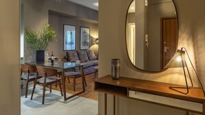 A modern living room with a glass dining table, wooden chairs, a sofa, and a coffee table. There is a large mirror on the wall, a floor lamp, and a potted plant. - Finesse Luxury Flat (Athens)