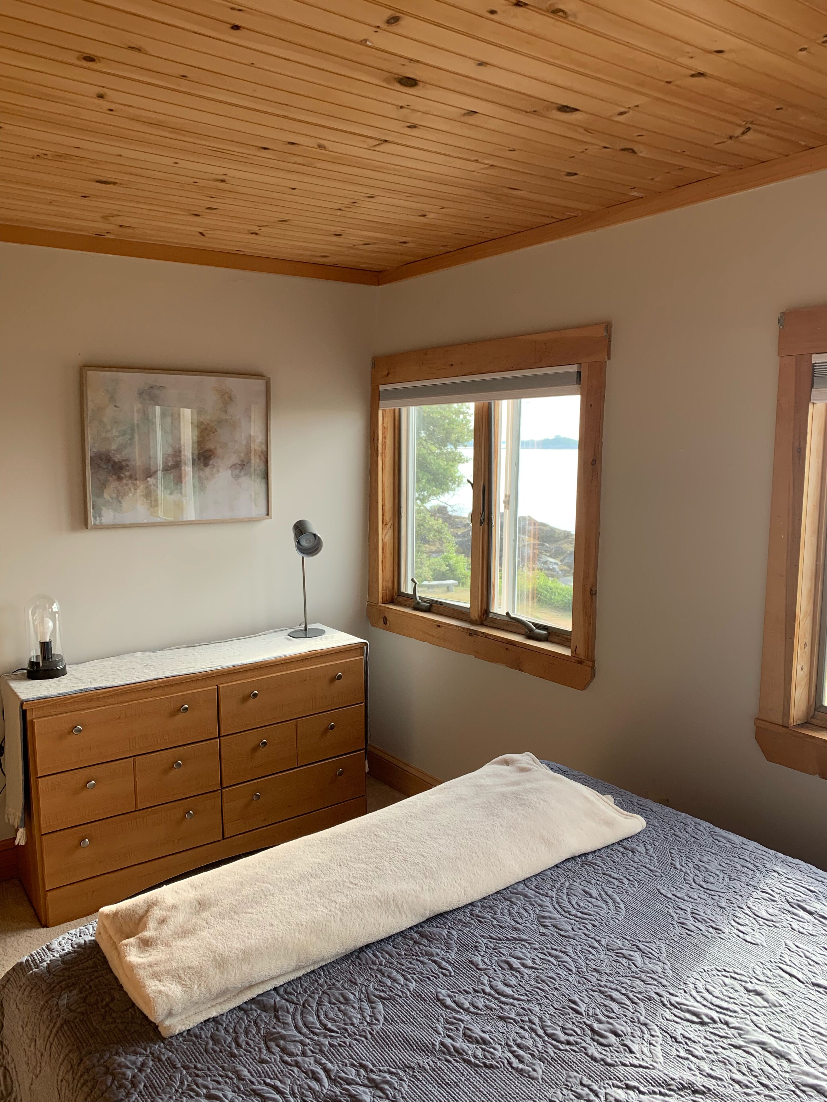 Un dormitorio con techo de madera, una cama con manta estampada, un aparador con lámpara y una ventana con vistas a la vegetación.