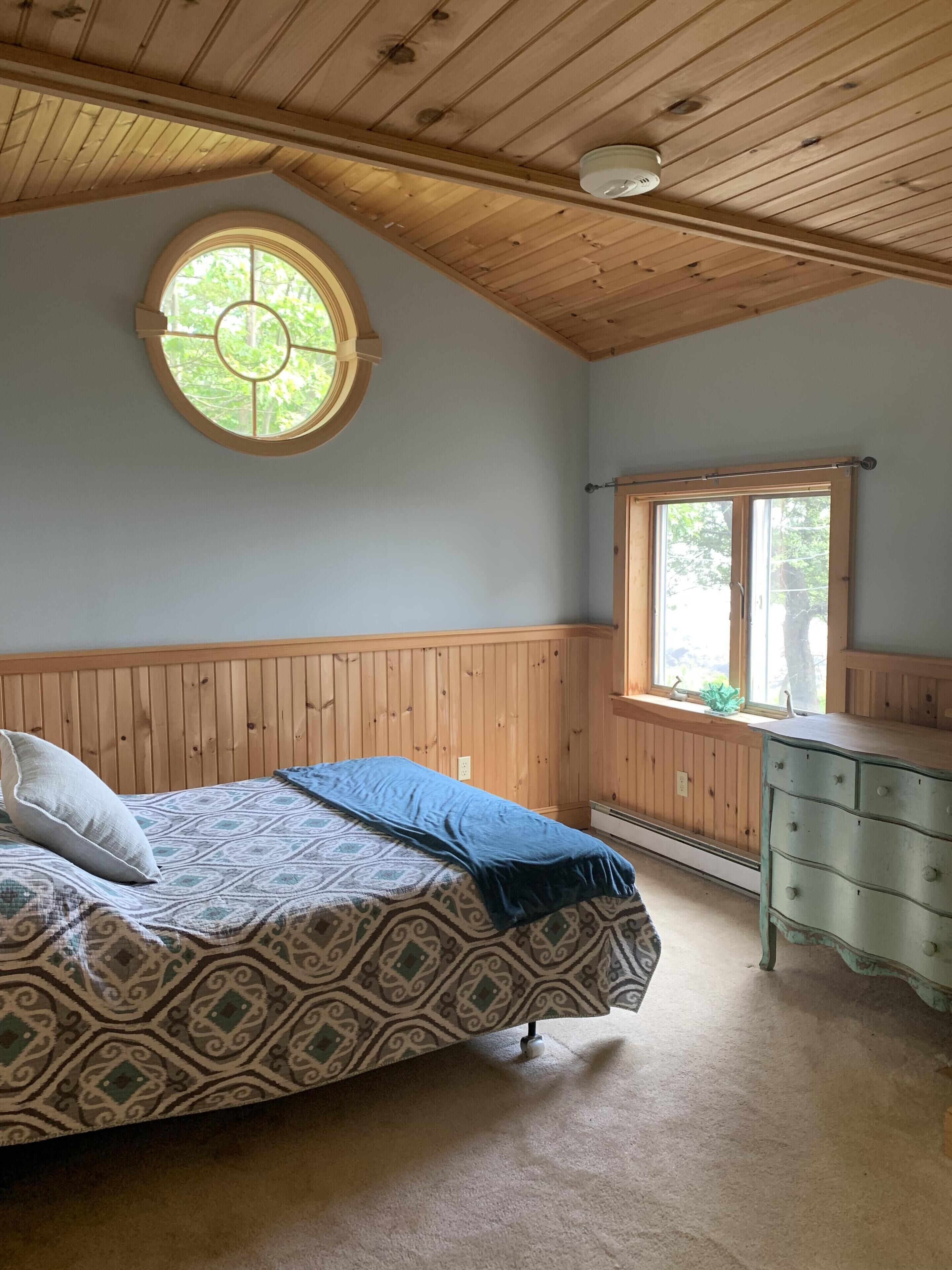 A bedroom with a wooden ceiling, a round window, a bed with patterned bedding, a dresser, and a window seat.
