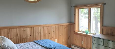 A bedroom with a wooden ceiling, a round window, a bed with patterned bedding, a dresser, and a window seat.