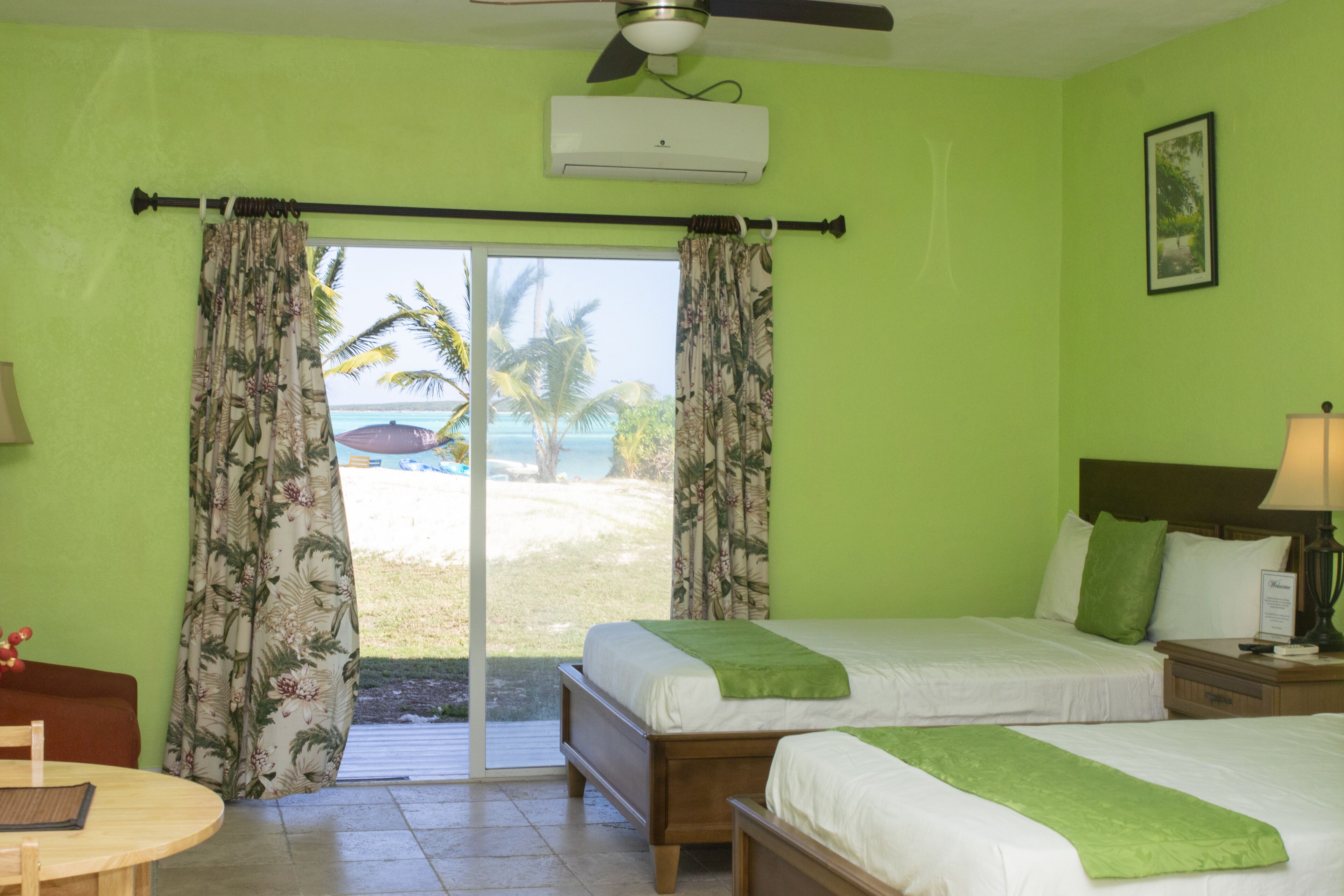 Hotelkamer met twee bedden, een plafondventilator, een airconditioning, een tafel met stoel, en uitzicht op het strand en palmbomen door het raam.