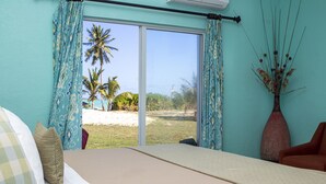 Quarto com cama, uma cadeira, um vaso com plantas secas e vista para palmeiras e uma praia através de uma porta de correr.